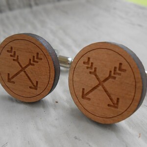 May include: A pair of wooden cufflinks with a circular design featuring two crossed arrows. The arrows are engraved into the wood and have a simple, minimalist style.