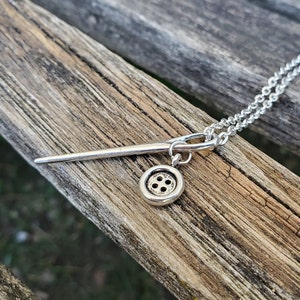 May include: A silver necklace with a needle and button charm. The needle is long and thin, and the button is round with four holes.