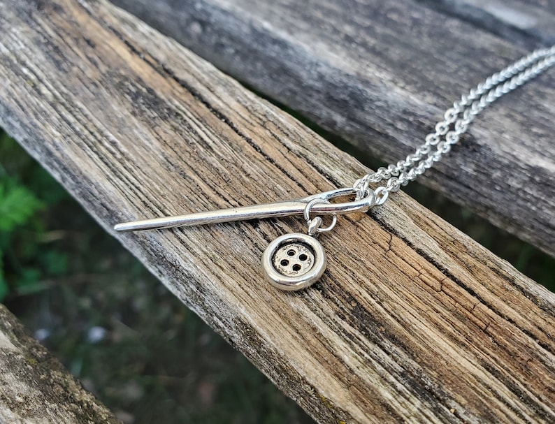 May include: A silver necklace with a needle and button charm. The needle is long and thin, and the button is round with four holes.