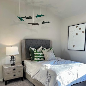 May include: A bedroom with a bed, nightstand, and artwork. The bed has a gray upholstered headboard, white bedding with paper airplane designs, and green and gray pillows. Above the bed, paper airplane mobiles hang from the ceiling. A framed print with paper airplane art hangs on the wall.