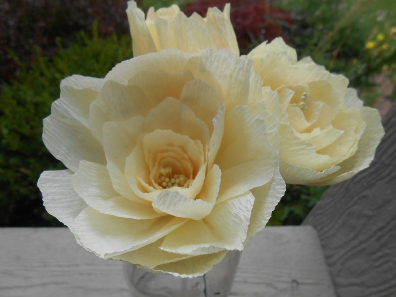 May include: Three cream-colored paper flowers in a glass vase. The flowers are made of crinkled paper and have a delicate, romantic look.