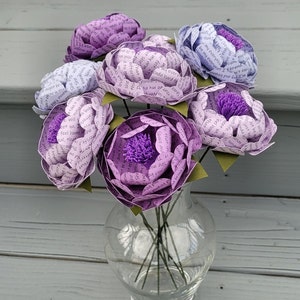 May include: A bouquet of six paper flowers in a clear glass vase. The flowers are made of paper with text printed on them. The flowers are in shades of purple and lavender.