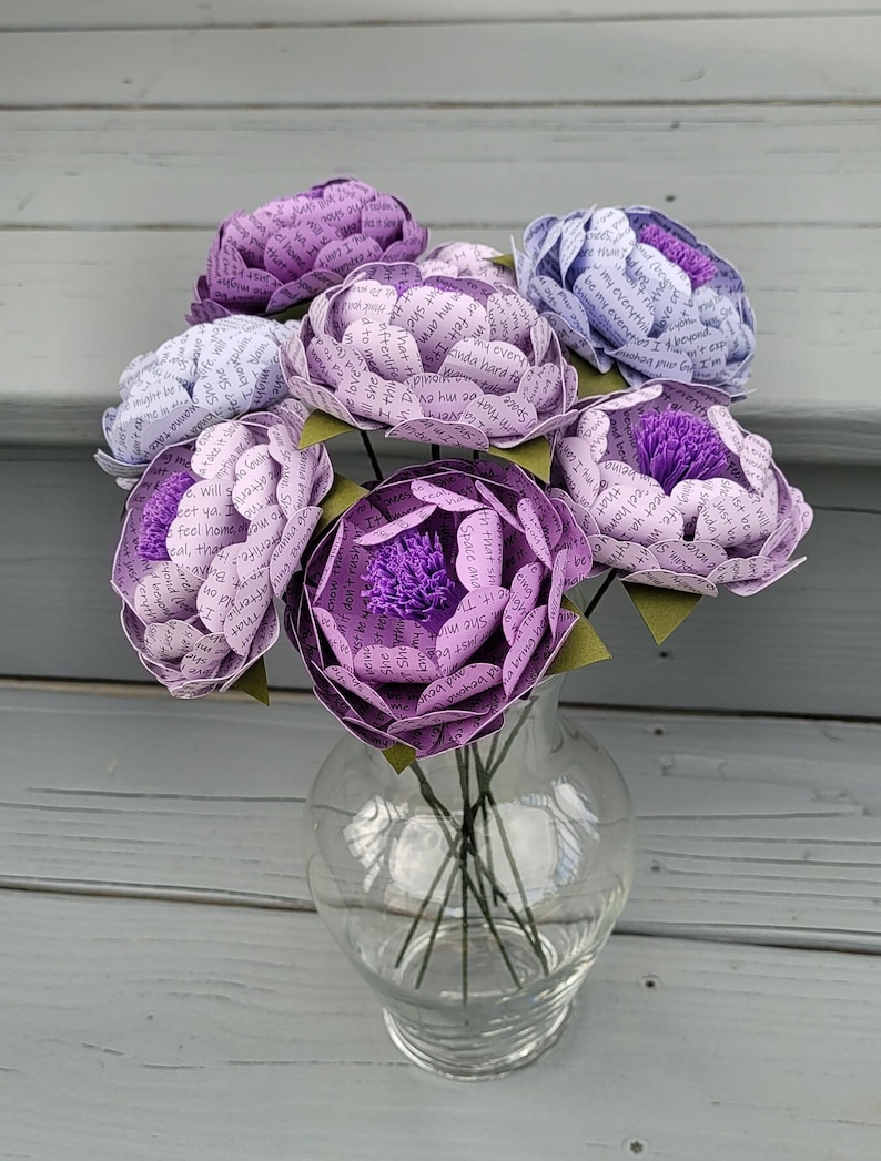 May include: A bouquet of six paper flowers in a clear glass vase. The flowers are made of paper with text printed on them. The flowers are in shades of purple and lavender.