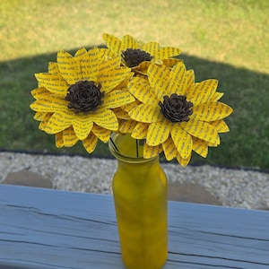 May include: Three paper sunflowers in a yellow vase. The sunflowers are made of paper with text printed on them. The vase is on a wooden surface.