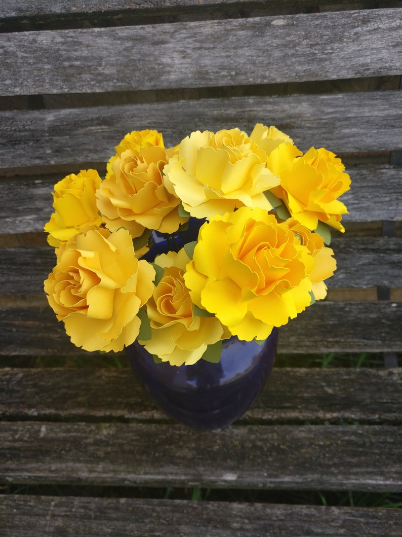 May include: A bouquet of yellow paper flowers in a blue vase. The flowers are arranged in a circular shape and have a realistic appearance.