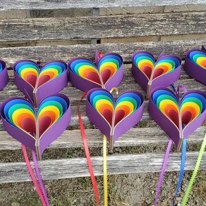 May include: Ten rainbow heart-shaped paper decorations with purple backs, hanging from a wooden bench. Each heart is made of layered paper in shades of red, orange, yellow, green, blue, indigo, and violet.