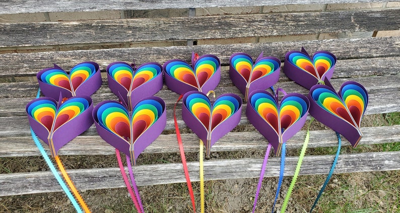 May include: Ten rainbow heart-shaped paper decorations with purple backs, hanging from a wooden bench. Each heart is made of layered paper in shades of red, orange, yellow, green, blue, indigo, and violet.