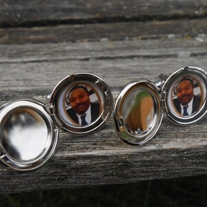 May include: Silver cufflinks with a round, hinged locket design. Each locket features a photo of a person with a dark complexion wearing a suit and tie.