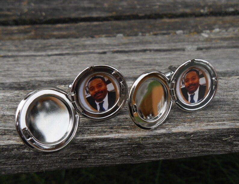 May include: Silver cufflinks with a round, hinged locket design. Each locket features a photo of a person with a dark complexion wearing a suit and tie.