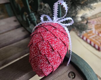 Könnte beinhalten: Ein roter Weihnachtsbaumschmuck aus Stoff mit weißem Sternmuster, geformt wie ein Tannenzapfen. Er hat ein weißes Band zum Aufhängen. Der Schmuck liegt auf einer Holzoberfläche. Eine festliche Dekoration.