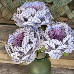 May include: Three paper flowers in a green vase. The flowers are made from white paper with purple centers and are decorated with printed text.
