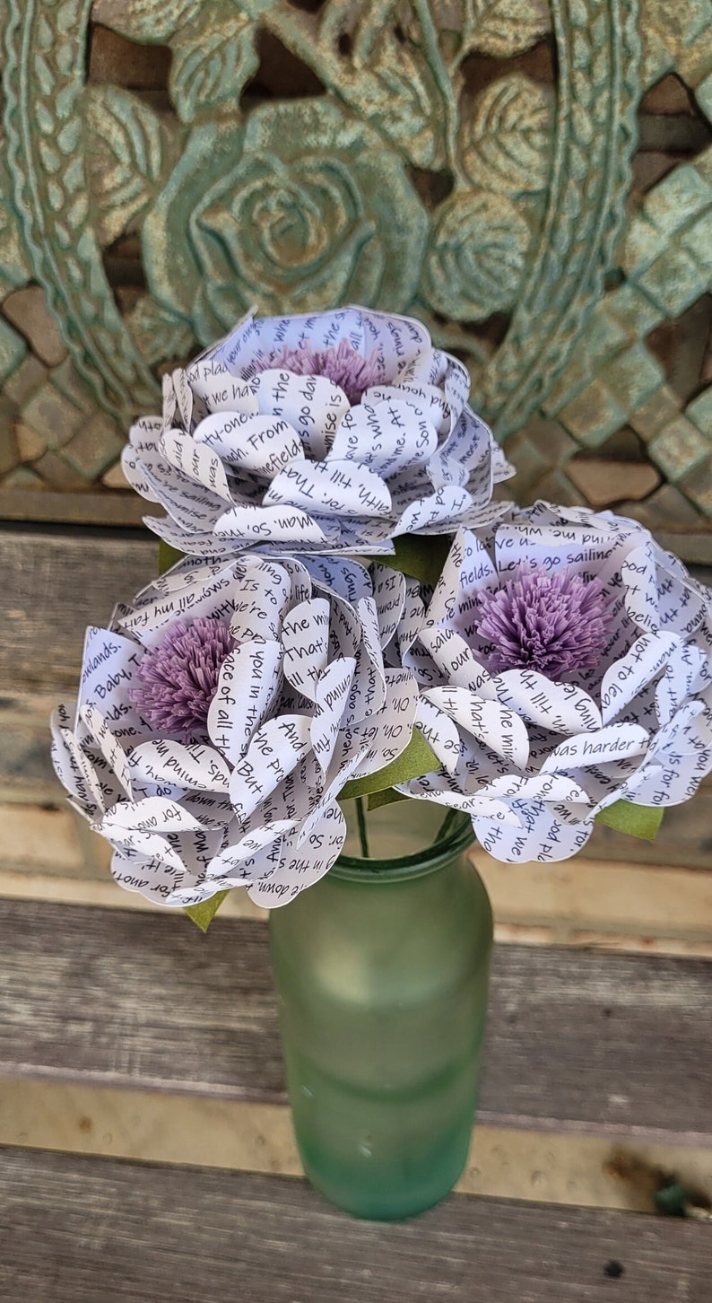 May include: Three paper flowers in a green vase. The flowers are made from white paper with purple centers and are decorated with printed text.