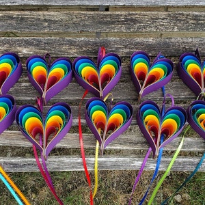 May include: Twelve rainbow paper hearts with purple centers and ribbon tails. The hearts are arranged in two rows of six on a wooden surface.
