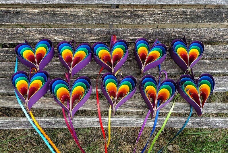 May include: Twelve rainbow paper hearts with purple centers and ribbon tails. The hearts are arranged in two rows of six on a wooden surface.