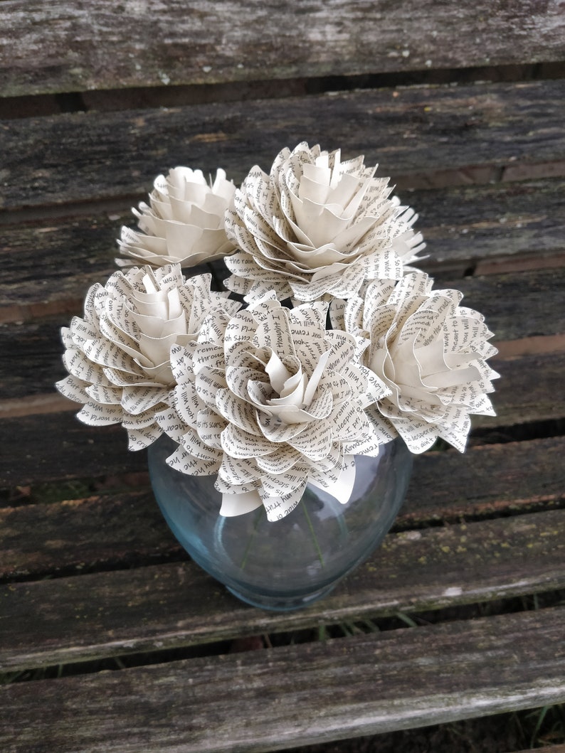 May include: A bouquet of paper flowers made from old book pages in a clear glass vase. The flowers are white and have a delicate, vintage look.