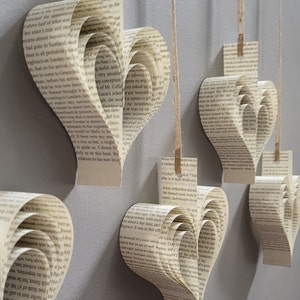 May include: Five paper hearts hanging from strings against a light gray wall. Each heart is made from pages of a book, with the text visible. The hearts are all different sizes and are arranged in a staggered pattern.