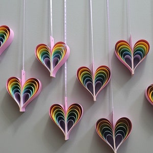 K&ouml;nnte beinhalten: Neun Regenbogen-Papierherzen h&auml;ngen an wei&szlig;en Schn&uuml;ren an einer hellgrauen Wand. Jedes Herz besteht aus mehreren Schichten Papier in Regenbogenfarben.