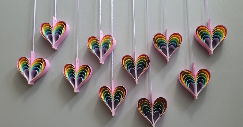 K&ouml;nnte beinhalten: Neun Regenbogen-Papierherzen h&auml;ngen an wei&szlig;en Schn&uuml;ren an einer hellgrauen Wand. Jedes Herz besteht aus mehreren Schichten Papier in Regenbogenfarben.