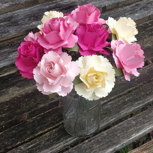 May include: A bouquet of paper flowers in shades of pink, yellow, and white arranged in a clear glass jar. The flowers are made of paper and have a realistic appearance.