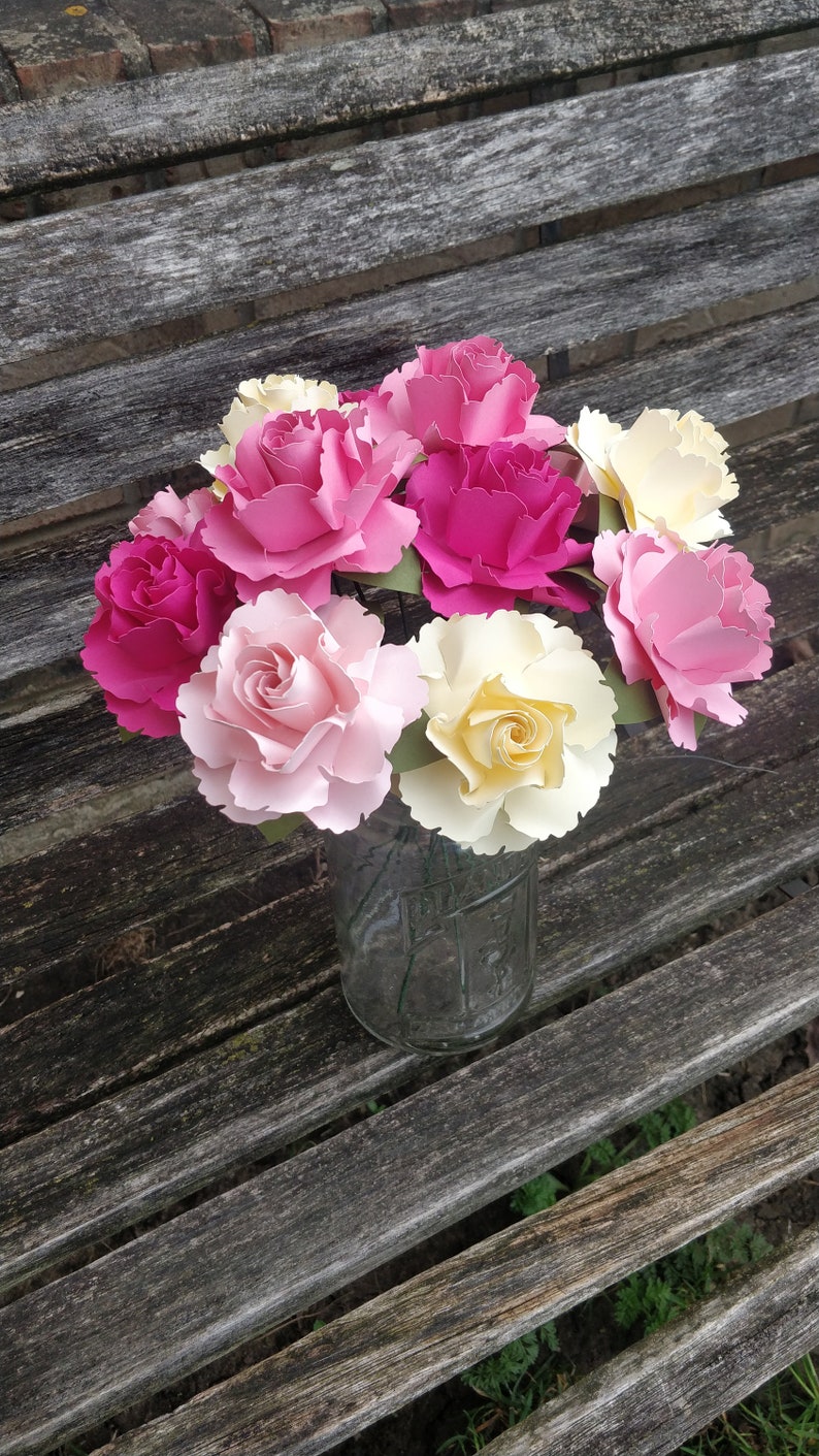 May include: A bouquet of paper flowers in shades of pink, yellow, and white arranged in a clear glass jar. The flowers are made of paper and have a realistic appearance.