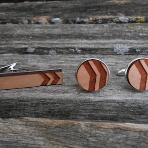 May include: A set of wooden cufflinks and a tie bar with a chevron pattern. The cufflinks are round and have a silver metal backing. The tie bar is rectangular and has a silver metal clip.
