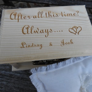 Puede incluir: Una caja de madera de color claro con tapa con bisagras. La tapa está grabada con las palabras "After all this time? Always...." seguidas de dos corazones y los nombres "Lindsay & Josh". Una almohada blanca con flecos está en primer plano.