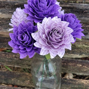 May include: A bouquet of purple and white paper roses made from book pages. The roses are arranged in a clear glass vase. The text on the pages is visible and includes phrases like "you shine, I", "our face", and "There are".