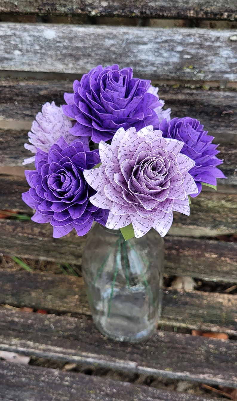 May include: A bouquet of purple and white paper roses made from book pages. The roses are arranged in a clear glass vase. The text on the pages is visible and includes phrases like "you shine, I", "our face", and "There are".