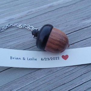 May include: A wooden acorn necklace with a silver chain. The acorn is dark brown with a black top. A white tag with the text "Brian & Leslie 6/23/2022" and a red heart is next to the necklace.