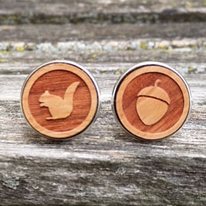 Puede incluir: Un par de gemelos de madera con un borde de metal plateado. Un gemelo presenta un diseño de ardilla, mientras que el otro presenta un diseño de bellota.