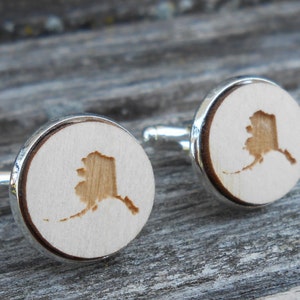 Peut inclure: Une paire de boutons de manchette en argent avec un incrustation en bois représentant le contour de l'état d'Alaska. Les boutons de manchette sont sur une surface en bois.