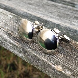 May include: A pair of silver cufflinks with round, smooth, reflective surfaces. The cufflinks are on a wooden surface.