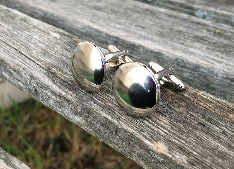 May include: A pair of silver cufflinks with round, smooth, reflective surfaces. The cufflinks are on a wooden surface.