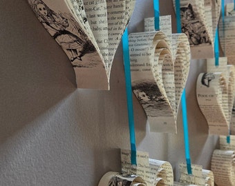 May include: A wall decorated with several book page hearts hanging from blue ribbon. The hearts are made from pages of books with text visible. The text includes "Alice in Wonderland" and "Pooh or T".