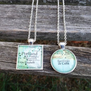 May include: Two silver necklaces with map pendants. The first pendant features a map of Portland, Maine, with the text "Portland" and "Cape Elizabeth". The second pendant features a map of Santiago de Cuba, with the text "Santiago de Cuba".