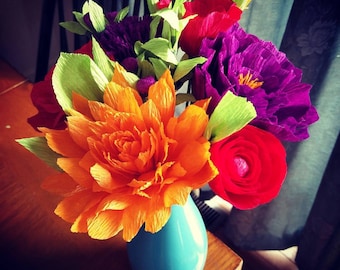May include: A bouquet of colorful paper flowers in a blue vase. The flowers are orange, red, purple, and green. The vase is a light blue color and has a round shape.