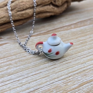 May include: A silver chain necklace with a small white teapot pendant. The teapot has red accents and a floral design.