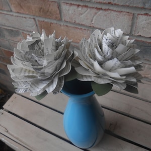 Pode incluir: Duas flores de papel branco com texto impresso são dispostas em um vaso azul claro. O vaso está sobre uma superfície de madeira branca.