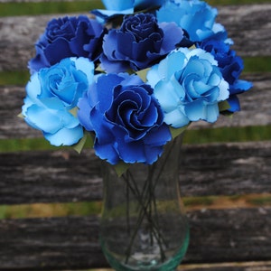 May include: A bouquet of blue and light blue paper flowers in a clear glass vase. The flowers are arranged in a circular pattern and have a delicate, layered appearance.
