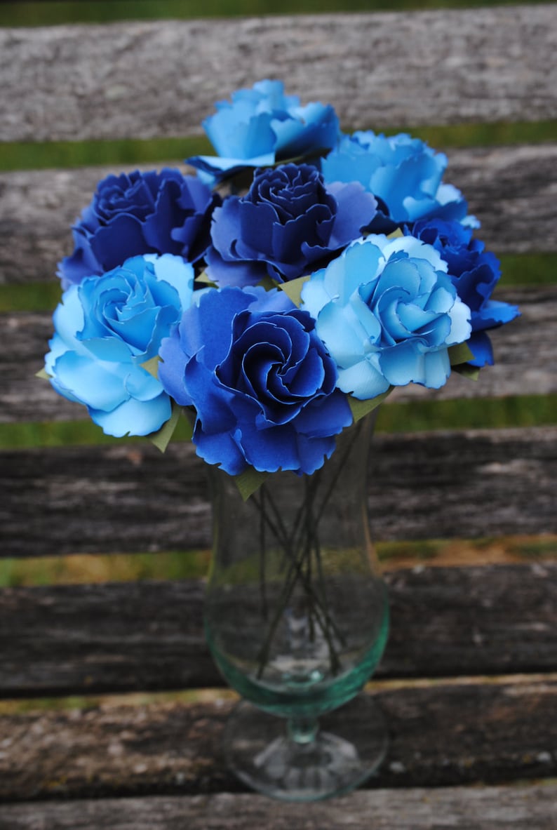 May include: A bouquet of blue and light blue paper flowers in a clear glass vase. The flowers are arranged in a circular pattern and have a delicate, layered appearance.