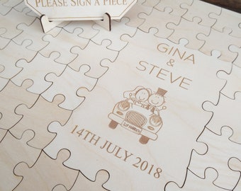 May include: A wooden guest book shaped like a puzzle with a sign that says "Guest Book Please Sign a Piece". The puzzle has a cartoon image of a couple in a car with the text "Gina & Steve 14th July 2018".