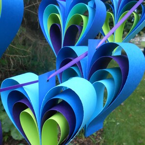 Pu&ograve; includere: Una ghirlanda di cuori di carta in tonalit&agrave; di blu, verde e viola. I cuori sono sovrapposti e piegati per creare un effetto tridimensionale.