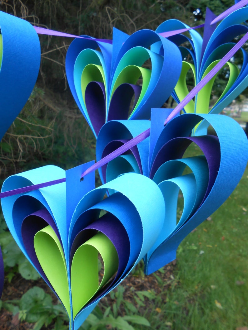Pu&ograve; includere: Una ghirlanda di cuori di carta in tonalit&agrave; di blu, verde e viola. I cuori sono sovrapposti e piegati per creare un effetto tridimensionale.