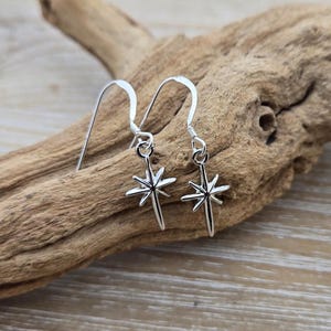 May include: A pair of silver-colored starburst earrings. Each earring features a star-shaped charm suspended from a hook. The earrings are displayed on a piece of driftwood.
