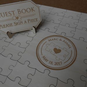 Könnte beinhalten: Ein Gästebuch aus Holz in Form eines Puzzles mit einem Schild mit der Aufschrift "Guest Book Please Sign a Piece". Das Puzzle hat die Form eines Herzens und ist mit den Namen "Marc & Ayana" und dem Datum "May 18, 2017" graviert.