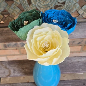 May include: Three paper flowers in a blue vase. The flowers are yellow, green, and blue. Each flower has a center made of white beads.