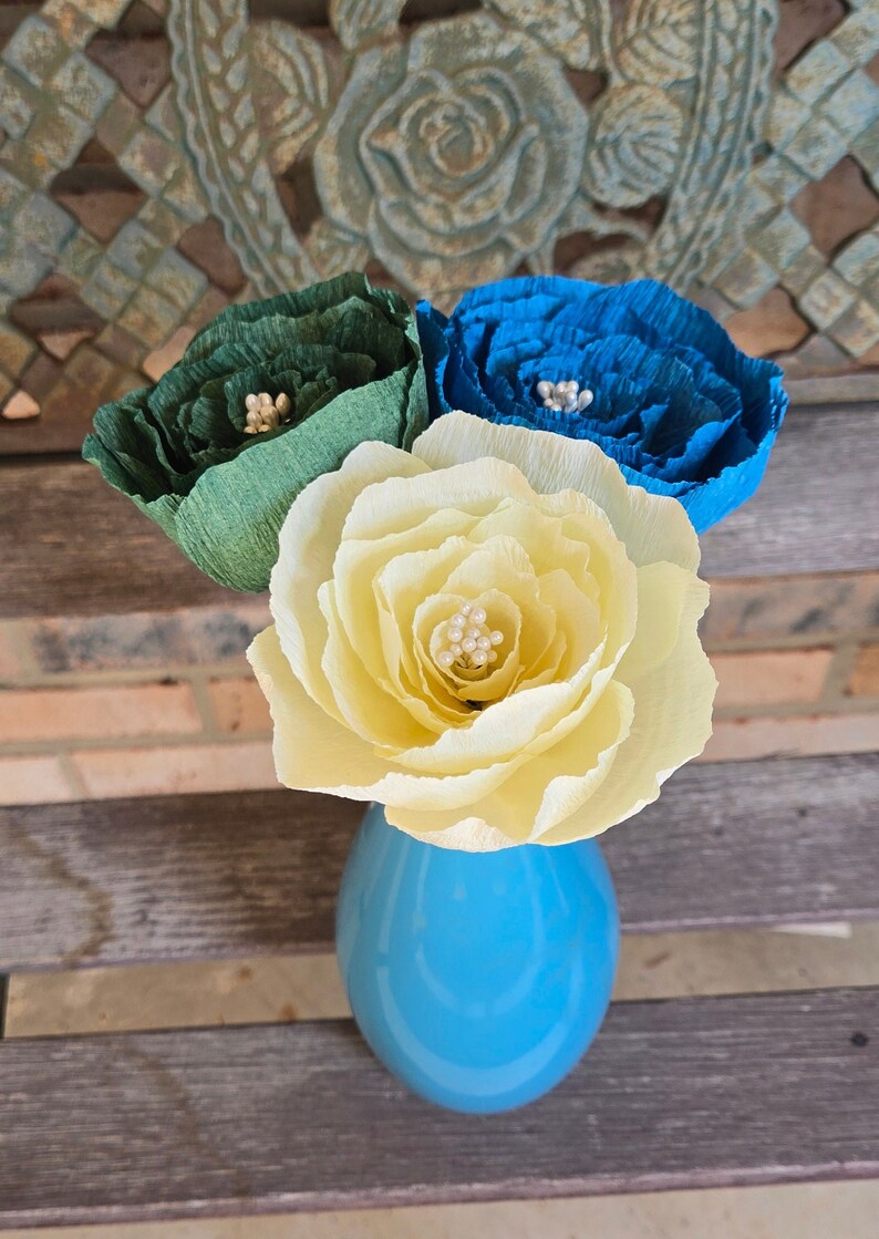 May include: Three paper flowers in a blue vase. The flowers are yellow, green, and blue. Each flower has a center made of white beads.