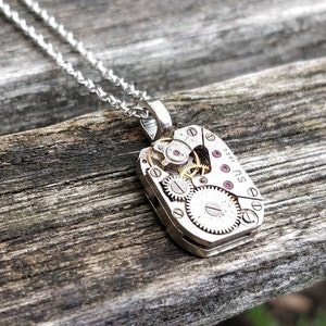 May include: A silver necklace with a pendant featuring a vintage watch movement with exposed gears and ruby jewels. The pendant is shaped like a rectangle and has the text "SU 001" on the side.