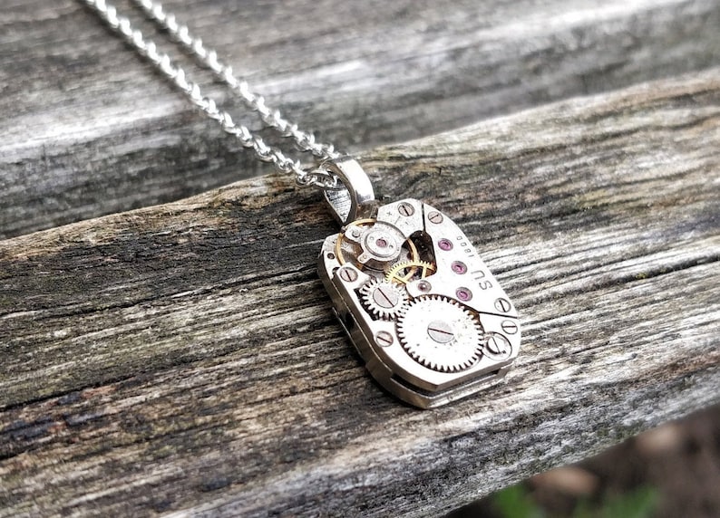 May include: A silver necklace with a pendant featuring a vintage watch movement with exposed gears and ruby jewels. The pendant is shaped like a rectangle and has the text "SU 001" on the side.