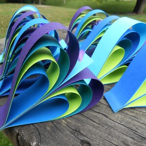 Pu&ograve; includere: Un gruppo di cuori di carta fatti di carta blu, verde e viola. I cuori sono piegati in modo da creare un effetto a strati.